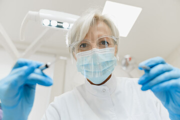 Low angle view of a caucasian middle-aged female dentist orthodontist stomatologist operating filling cleaning tooth teeth mouth cavity in protective glasses