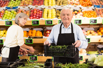 Elderly supermarket employee rearranges a box of green chili peppers