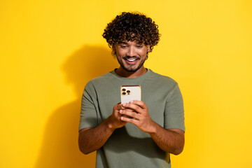 Wall Mural - Photo of smart positive guy with wavy hairdo dressed khaki t-shirt look at smartphone typing message isolated on yellow color background