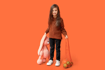 Wall Mural - Cute stylish little girl holding string bag with pumpkins and bunny toy on orange background