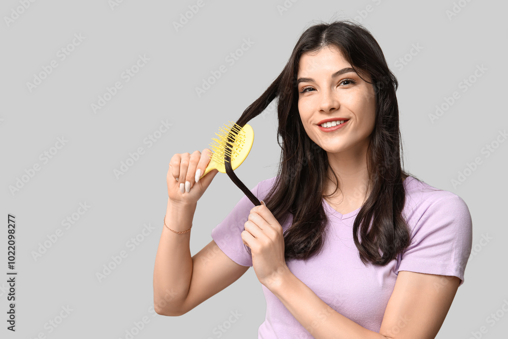 Beautiful young woman brushing her hair on grey background