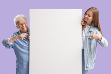 Wall Mural - Happy senior woman and her granddaughter pointing at blank poster on purple background