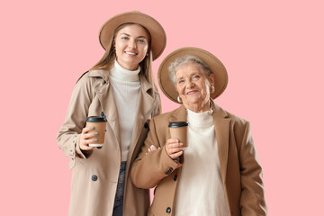 Wall Mural - Happy senior woman and her granddaughter in autumn outfits with cups of coffee on pink background