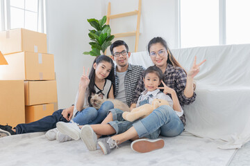 Happy Asian family with two children and cat sitting on couch in new home, surrounded by moving boxes. Parents and kids making peace signs. cardboard boxes, playfully unpacking. Relocation concept.