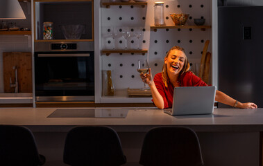 Smiling woman working at home using laptop while drinking wine at night.