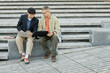 © Seventyfour - Full shot of Asian male IT developer using laptop collaborating with colleague on work while writing code sitting on concrete bench outside in city, copy space