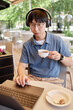 © Seventyfour - Vertical shot of Asian male IT developer in headphones ang glasses keeping fingers on touchpad using laptop to work while holding cup of coffee at table in outdoor seating area of cafe, copy space