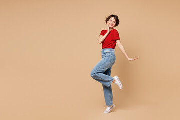 Full body young smiling happy woman she wears red t-shirt casual clothes raise up leg put hand on face look camera isolated on plain pastel light beige background studio portrait. Lifestyle concept.