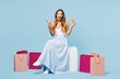 © ViDi Studio - Full body young woman wear casual clothes sits near paper package bags using mobile cell phone credit bank card do online shopping isolated on plain blue background. Black Friday sale buy day concept.