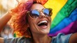 © Dulemegapixel - A joyous woman with vibrant hair and sunglasses beams broadly against a rainbow pride flag, symbolizing self-expression and celebration of diversity.