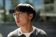 © Seventyfour - Close up of adult Asian man with glasses looking away lost in thought while standing outside in sunlight on blurred background, copy space