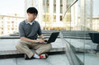 © Seventyfour - Serious Asian man in stylish gray polo outfit concentrated on using laptop typing on keyboard while sitting on concrete steps outside in city street, copy space