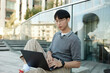 © Seventyfour - Medium shot of Asian male IT programmer typing on laptop sitting on sidewalk leaning on glass construction while working in public space in city street, copy space