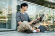 © Seventyfour - Medium shot of Asian male IT programmer looking at laptop screen keeping fingers on touchpad while sitting on sidewalk next to glass construction working in public space in city street, copy space