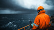 © Marketplace - A worker in an orange safety suit stands on a ship, observing dark storm clouds over turbulent waters, preparing for challenging maritime conditions.