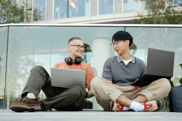 Candid shot of two smiling IT programmers from different ethnic backgrounds having fun while working on laptops collaboratively sitting on pavement outside in city street, copy space