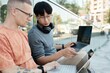 © Seventyfour - Medium shot of Asian businessman with headphones pointing at laptop screen working together with colleague on project while sitting on pavement in city street, copy space