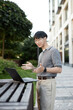 © Seventyfour - Vertical shot of smiling Asian businessman looking at laptop screen talking with colleague captured during online conference while working at stand up desk outside in city street, copy space