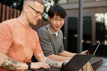 Candid shot of smiling Asian male IT programmer working on laptop collaboratively with colleague having fun while sitting on bench outside in city street, copy space