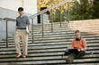 © Seventyfour - Wide angle shot of Asian businessman in office clothing walking down stairs where male colleague using laptop to work sitting on steps outside in public space in city, copy space