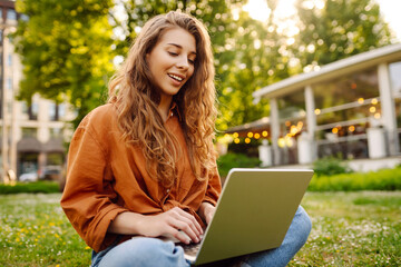 Wall Mural - Happy woman with a laptop sitting on a green meadow. Education online. Freelance work, technology concept.