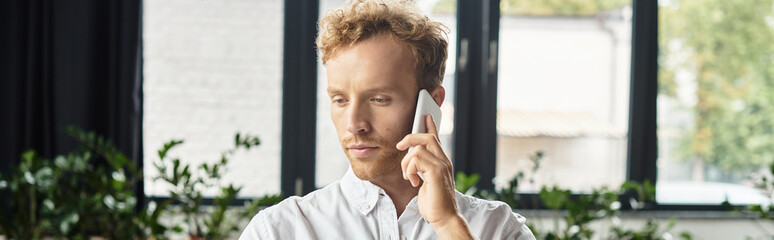 A focused redhead businessman discusses project details over the phone in a bright office.