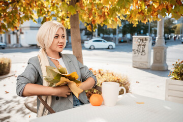 Wall Mural - A blonde woman sits at a table with a cup of coffee and a stack of papers. She is wearing a gray jacket and a necklace. The scene is set in a park with trees and benches.