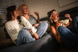 © BalanceFormCreative - A  mother and her two daughters sit together on the couch at night, watching an intense TV show. They are focused on the screen while sharing a big bowl of popcorn.
