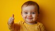 © weerayut - A joyful baby wearing a yellow outfit gives a thumbs up against a vibrant yellow background, exuding positivity and happiness.