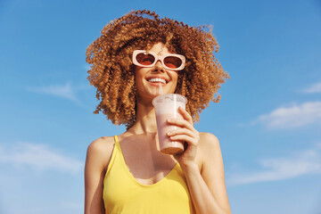 Happy woman with curly hair holding a smoothie on a sunny day against a blue sky