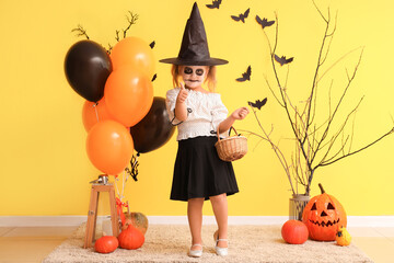 Wall Mural - Little girl dressed for Halloween as witch with candies showing thumb-up near yellow wall