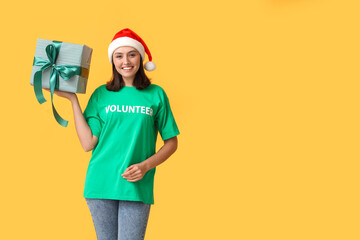 Wall Mural - Beautiful female volunteer in Santa hat with gift box on yellow background