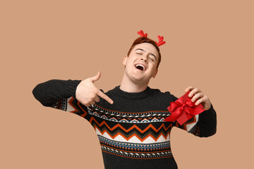 Poster - Handsome young man in warm sweater and reindeer headband pointing at gift box on beige background