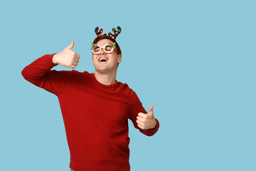 Poster - Handsome young man in warm sweater, reindeer headband and novelty glasses showing thumbs-up on blue background