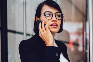 Concentrated financial manager in elegant wear talking on smartphone near business center, serious woman trader in eyeglasses having phone conversation about startup project during coffee time