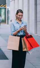 Wall Mural - cheerful female shopaholic enjoying time for shopping in downtown typing message to friend about bla