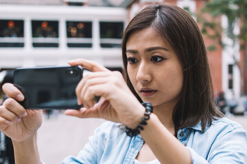 Japanese young woman tourist focusing mobile camera on city urban space and making pictures on modern smartphone device to uploading photos in internet blog standing outdoors in historic center