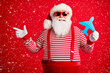 © deagreez - Photo of grandfather grey beard hold blue paper plane direct finger empty space wear santa claus x-mas costume suspenders sunglass striped shirt cap isolated red color background