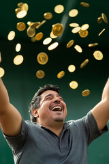 Middle-aged Hispanic man in football stands, arms raised high, laughing in surprise as gold coins fly through the air.