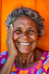 Wall Mural - Senior Brazilian woman with a big, heartfelt smile, wiping away a tear of gratitude, her eyes full of appreciation, set against a vibrant orange background.