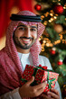 © wolfhound911 - Young adult Arabian man smiling and holding a Christmas present, standing by a festive tree, red background.