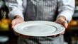 © Svetlana - Waiter wearing an apron, holding an empty plate with both hands in a restaurant. Hospitality and service concept