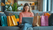 © tisomboon - A woman sits on a sofa with colorful shopping bags, smiling while using her laptop in a cozy living room.