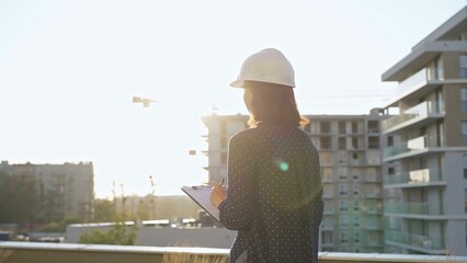 Female architect wearing a safety helmet is using a tablet computer while inspecting a building construction site at sunset