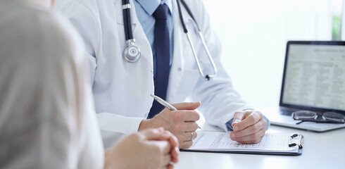 A doctor and a patient. The physician, wearing a white medical coat over a blue shirt and tie, is filling out a medical record form during a consultation in the clinic. Medical service