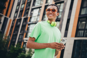 Wall Mural - Portrait of nice young man hold phone headphones hang neck wear t-shirt weekend walk downtown street outdoors