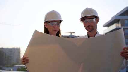 two architects colleagues, wearing safety helmets and glasses, are examining blueprints at a constru