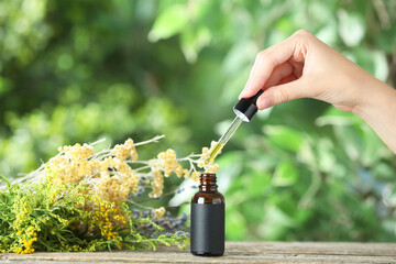 Wall Mural - Woman dripping tincture from pipette into bottle at wooden table against blurred green background, closeup