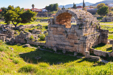 Wall Mural - Ancient ruins in the Corinth Archaeological Park, featuring a large stone structure, green grass, and surrounding trees under a bright blue sky. Corinth, Greece