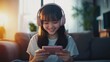 © Thavesak - Happy East Asian Teenage Girl Playing Phone Games in Sunlit Modern Living Room